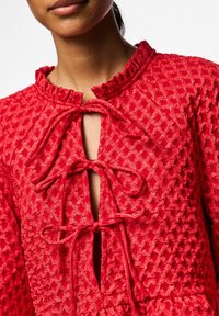 Sarkans teksturēts kleita ar ruļļošu dekoltē, atslēgas caurumu, kas nostiprināts ar saitēm, un sarežģītiem rakstiem visā audumā.