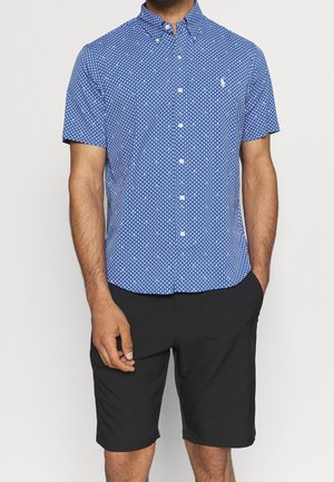 Homme portant une chemise bleue à manches courtes à motifs et un short noir mi-genoux, les mains dans les poches.