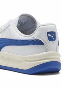Weißes Leder-Sneaker mit blauen Akzenten, ausgestattet mit einem strukturierten blauen Streifen, einem gepolsterten Kragen und einer strukturierten Gummisohle, die das Puma-Logo zeigt.