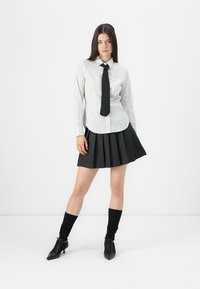 Young woman with long dark hair wearing a white striped shirt, black necktie, pleated black skirt, black socks, and black heeled shoes standing.
