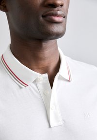 Hvitt polo skjorte med krage, prydet med røde og beige striper, knappeåpning og subtilt brodert logo på brystet. Glatt tekstur.