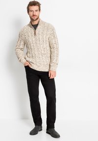 Maglione beige a trecce con zip a metà colletto, abbinato a pantaloni neri e stivaletti neri, mani nelle tasche, sfondo neutro.