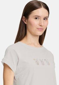 Lys grå t-shirt med rund hals, opkravede ærmer og et dekorativt perlemønster foran, der viser farverige designs.