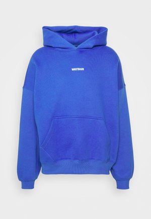 Personne portant un pantalon de survêtement bleu vif et un sweat-shirt assise sur une mallette en métal avec des baskets blanches sur un tapis bleu clair.