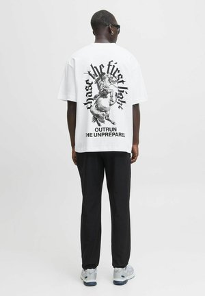 Mand med hvid grafik t-shirt med en kriger på en hest og teksten "forfølg det første lys" og "overhal den uforberedte," sorte bukser, sneakers.