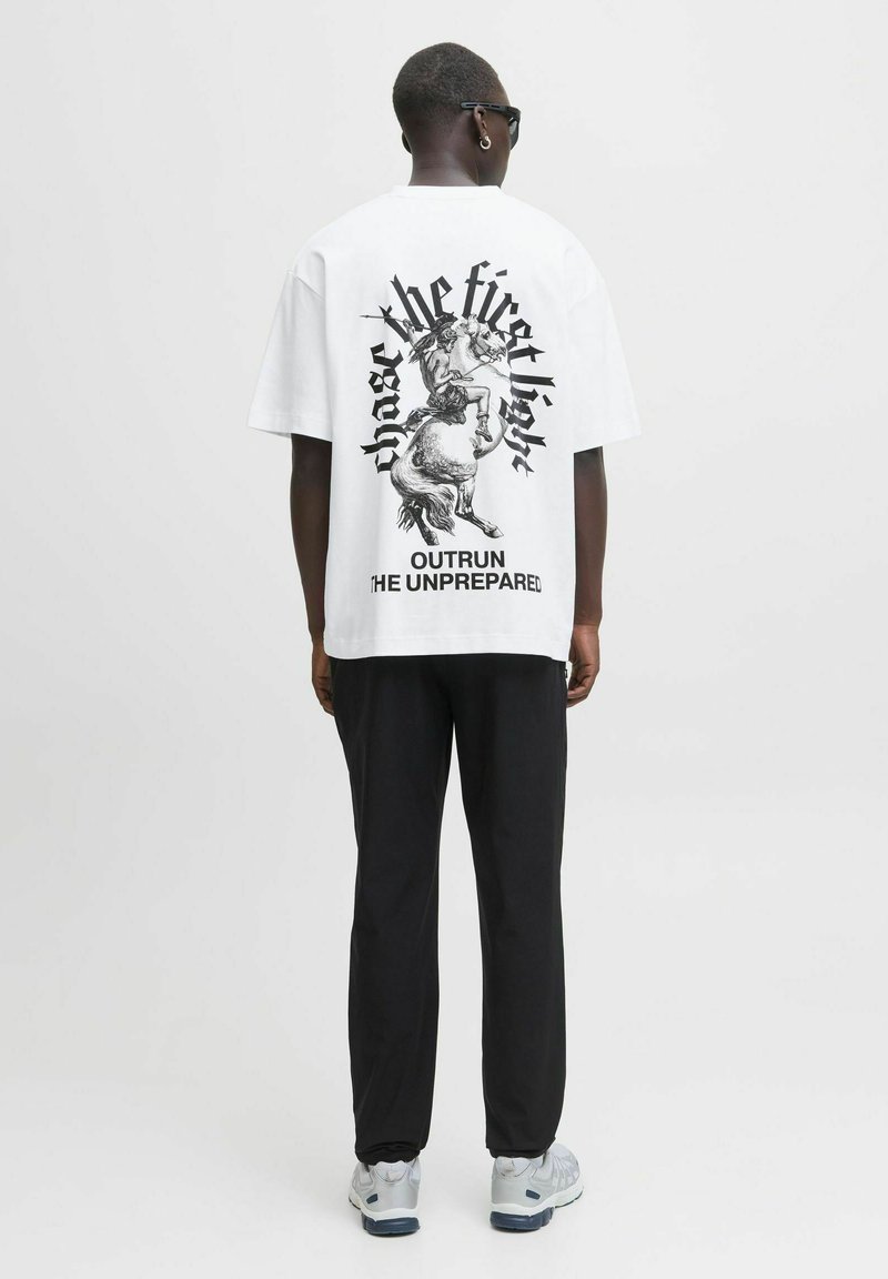Mand med hvid grafik t-shirt med en kriger på en hest og teksten "forfølg det første lys" og "overhal den uforberedte," sorte bukser, sneakers.