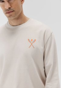 Sudadera de manga larga color beige claro con un gráfico naranja de utensilios cruzados y un logotipo de pulpo en el área del pecho izquierdo. Textura suave.