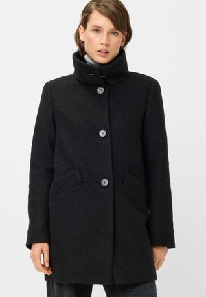 Woman wearing a black buttoned wool coat with high collar over a light gray turtleneck sweater, standing against a plain background.