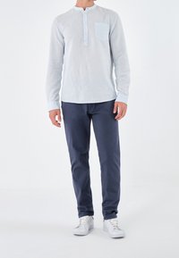 Man in een lichtblauw overhemd met lange mouwen, donkere blauwe broek en witte sneakers, staande tegen een effen witte achtergrond.