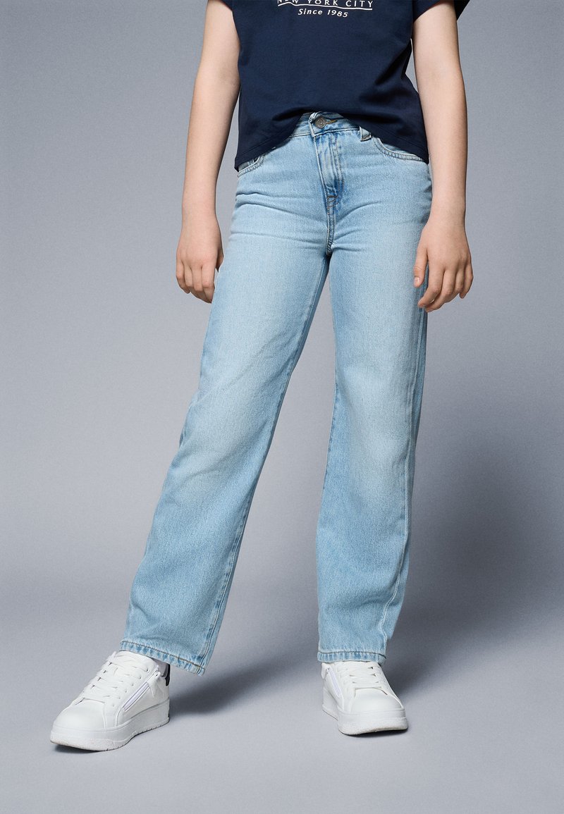 Light blue straight-leg jeans made of denim, featuring a mid-rise waist, slight fading, and a classic five-pocket design, paired with white sneakers.