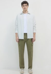 Jeune homme portant une chemise blanche à rayures avec un t-shirt blanc, un pantalon cargo vert olive et des chaussures beige clair, se tenant devant un fond blanc uni.
