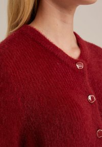 Sarkana adīta džemperis ar dziļu V-veida kakla izgriezumu un trim lielām, apaļām, spīdīgām pogām. Mīksta, pūkaina tekstūra. Vienkāršs, klasiskas dizains.
