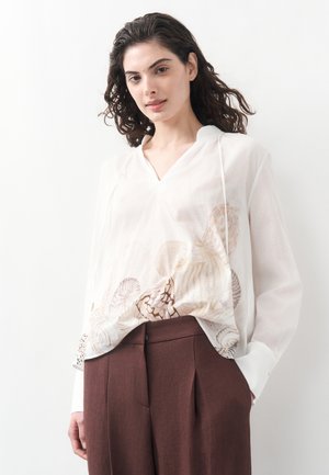 Young woman with curly hair wearing a white blouse with shell patterns and brown high-waisted trousers, standing against a white background.