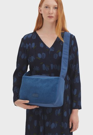 Borsa a spalla in velluto a coste blu e tessuto, con una parte frontale piatta, tracolla regolabile e forma rettangolare, tenuta da una persona che indossa un vestito a fantasia.