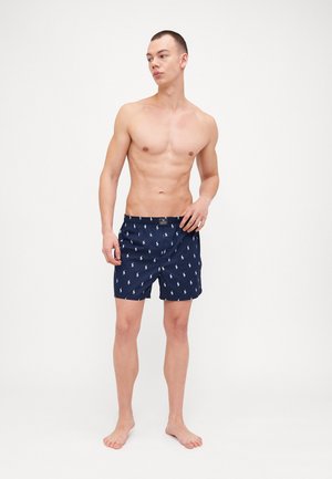 Shorts de bain navy avec un motif léger de petits logos blancs, taille élastique et coupe décontractée, fabriqués en matériau à séchage rapide.