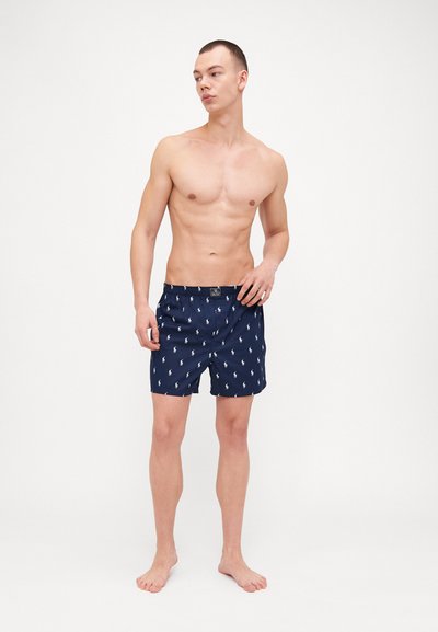 Marineblå svømshorts med et let mønster af små hvide logoer, elastisk talje og en afslappet pasform, lavet af hurtigtørrende materiale.