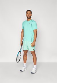 Tricou sportiv ușor, de culoare verde mentă, cu mâneci scurte și accente negre, asortat cu pantaloni scurți mint. Jucătorul �ține o rachetă de tenis și o minge.