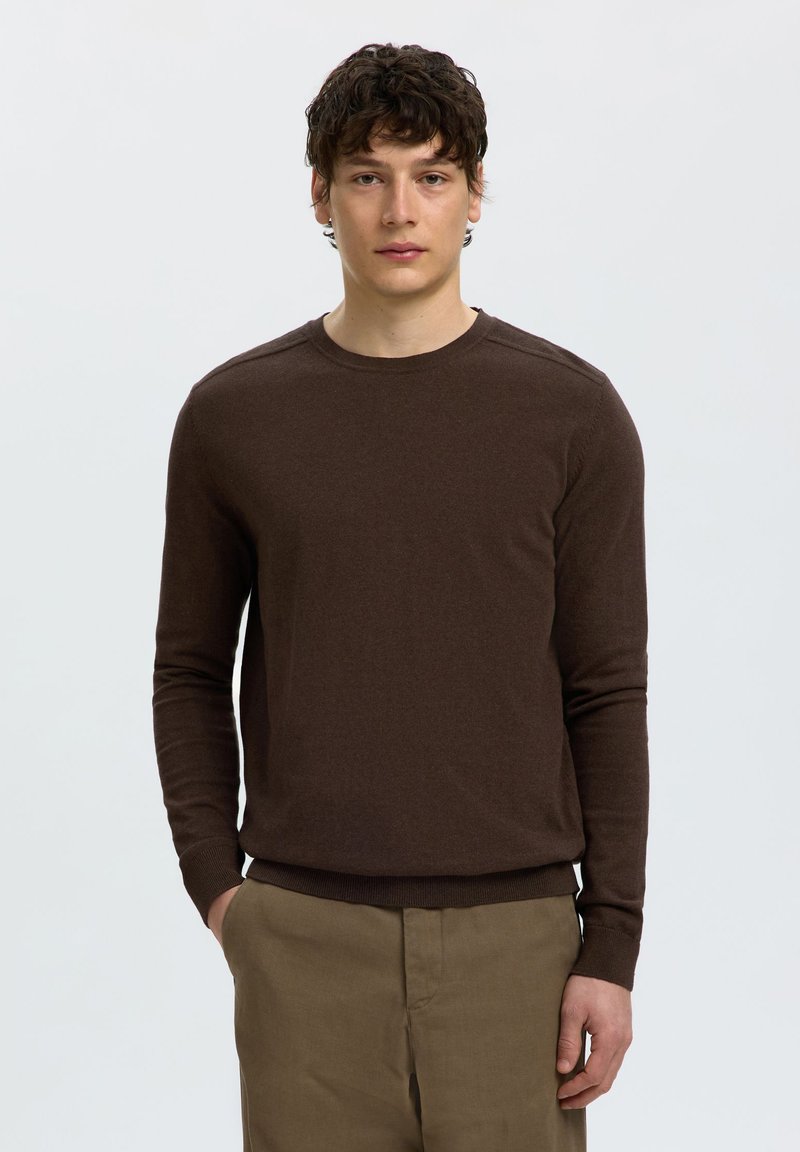 Jeune homme aux cheveux courts et bouclés portant un pull marron à manches longues et un pantalon kaki sur un fond clair uni.