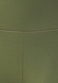 Materiał sportowy w oliwkowym kolorze, charakteryzujący się gładką teksturą, płaskimi szwami oraz subtelnymi detalami wzdłuż pasa i nogawek.