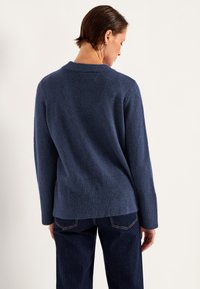 Dunkelblauer Pullover aus weichem Stoff, mit geripptem Saum und lässigem Schnitt, kombiniert mit dunklen Jeans; von hinten betrachtet.