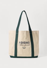 Beigā Adidas auduma soma ar tumši zaļiem rokturiem un apakšu, ar tekstu "Adidas Athletic Garment" un logo priekšpusē.