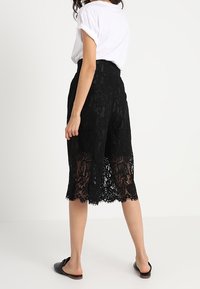 Černé krajkové culottes s poloprůhledným designem, vlněným lemem a vysokým pasem, spárované s bílým tričkem a černými plochými sandály.