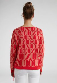 Czerwony sweter z dużym, powtarzającym się wzorem logo w różnych odcieniach czerwieni. Długie rękawy, luźny krój i okrągły dekolt.