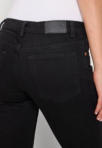 Vue de dos d'une personne portant un jean noir avec des poches arrière et une étiquette noire indiquant "Monki" sur la ceinture, associée à un haut blanc.