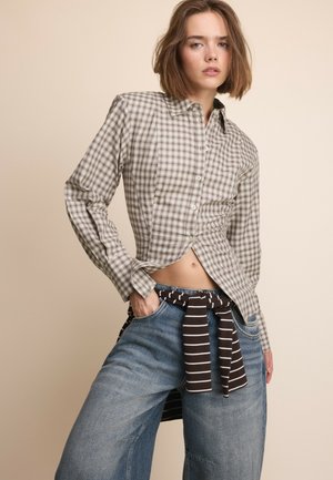Jeune femme debout portant une chemise à manches longues à carreaux, un short en jean large, des bottes noires et un pull rayé noué autour de la taille.