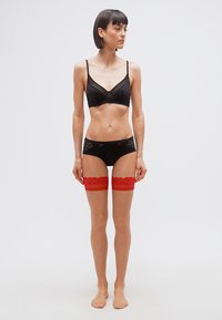 Γυναίκα με κοντά σκουρόχρωμα μαλλιά, όρθια και ξυπόλυτη, φοράει μαύρα εσώρουχα και κόκκινες δαντελωτές κάλτσες πάνω από το γόνατο, κοιτάζει προς τα αριστερά της.