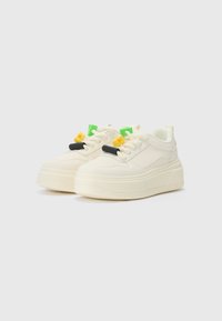 Weiße Sneakers mit einer klobigen Sohle, strukturiertem Obermaterial und kontrastierenden Akzenten in Grün, Gelb und Schwarz an den Schnürsenkeln.