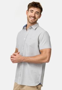 Camisa de manga corta en gris claro con cuello abotonado, que presenta un interior del cuello con un patrón contrastante y un sutil logo en el pecho.