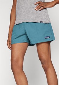 Teal shorts made from lightweight fabric, featuring an elastic waistband and side pockets, with a prominent brand logo on the hem.