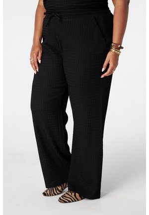 Femme portant un pantalon large noir texturé avec taille à cordon et des ballerines pointues imprimé tigre, mains le long du corps avec bagues et bracelets.