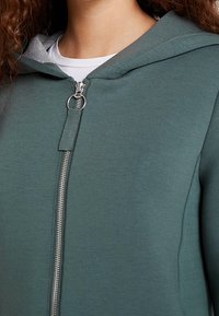Vrouw in een groene hoodie met rits en een zilveren ring aan de rits, met een wit shirt zichtbaar eronder.