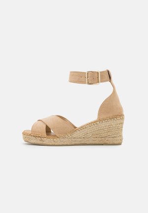 Sandalias de cuña de ante beige con una correa de tobillo ajustable, diseño de tiras cruzadas y suela de cuña envuelta en yute. Textura suave y color neutral.