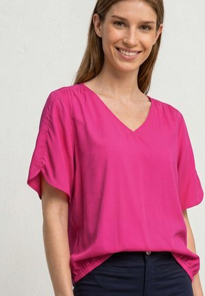 Femme souriante portant un chemisier ample rose vif à manches courtes et col en V, associé à un pantalon foncé, sur un fond uni clair.