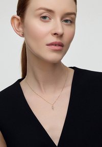 Złoty naszyjnik z delikatnym zawieszką w kształcie liścia, noszony przez modelkę o jasnej karnacji, na tle czarnej bluzki i neutralnego tła.