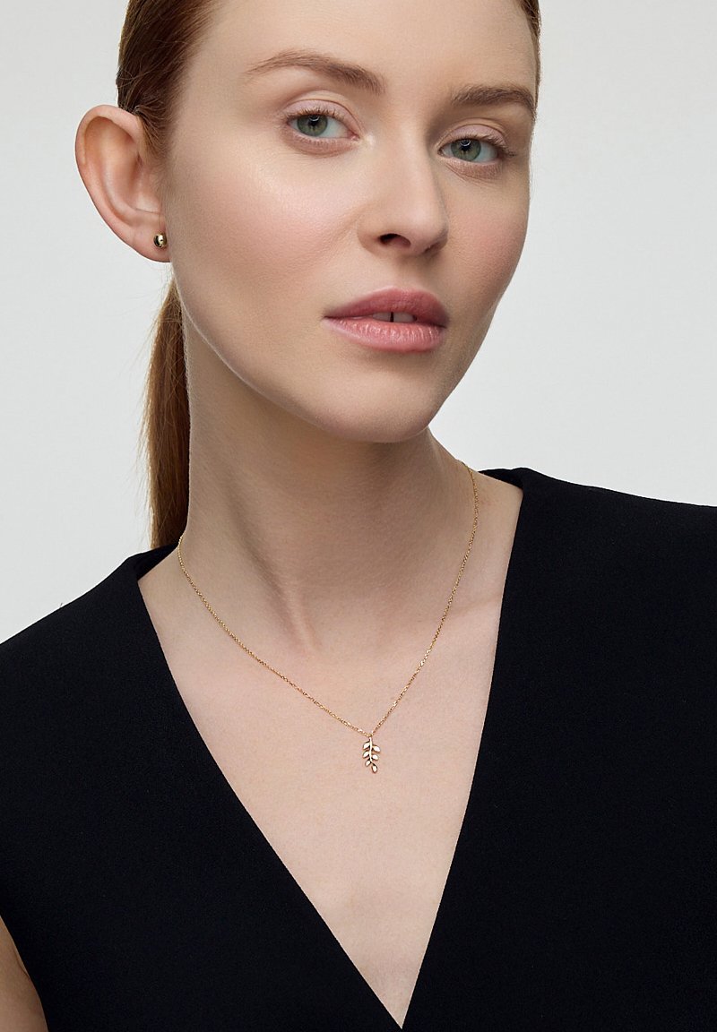 Złoty naszyjnik z delikatnym zawieszką w kształcie liścia, noszony przez modelkę o jasnej karnacji, na tle czarnej bluzki i neutralnego tła.