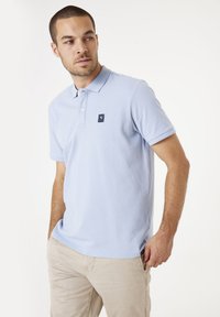 Lichtblauw poloshirt met een gestructureerde weving, een placket met twee knopen en een klein zwart logo op de linkerborst. Gecombineerd met beige broeken.