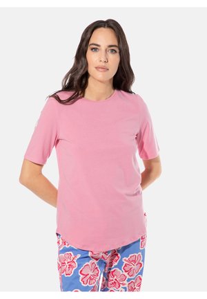 Frau mit langen dunklen Haaren, die ein rosa kurzärmeliges Hemd und blaue Hosen mit pinken Hibiskusblumenmustern trägt, stehend mit den Händen hinter dem Rücken.