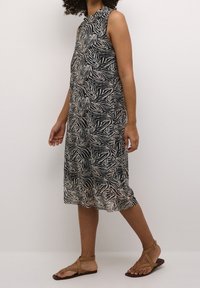 Femme portant une robe midi sans manches à motif zèbre noir et blanc et des sandales plates à lanières marron, debout devant un fond clair uni.