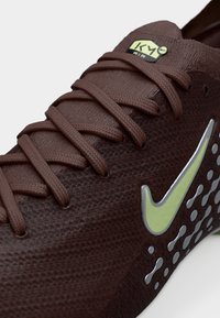 Brun sportsko laget av teksturert materiale. Har avrundet tå, lisser, en limegrønn Nike-swoosh og ventil-lignende perforeringer på siden.