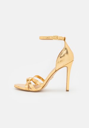 Sandale à talon doré avec une finition métallique texturée, une bride de cheville avec boucle, et un design à brides croisées sur l'avant-pied.