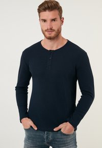 Mornarsko modra dolgi rokav henley majica z teksturirano tkanino, okroglo ovratnico in trojnim gumbom. V kombinaciji z svetlimi kavbojskimi hlačami.