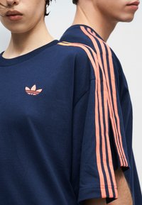 Δύο άτομα φοράνε μπλε ναυτικό πουκάμισα Adidas με ροζ τρεις ρίγες στο μανίκι και ροζ λογότυπο trefoil της Adidas στο στήθος.