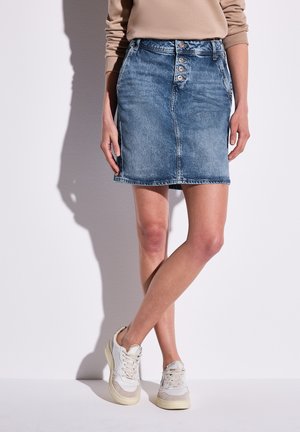 Jupe en denim bleu moyen au design ajusté, avec une fermeture à cinq boutons à l'avant et un ourlet brut. Assortie à des baskets blanches avec des accents beiges.