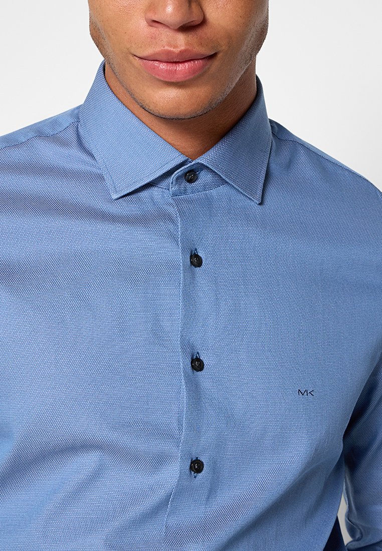 Chemise bouton-brisée bleu clair avec un tissu texturé ; elle présente un col étroit, des boutons noirs et un petit logo brodé sur la poitrine.
