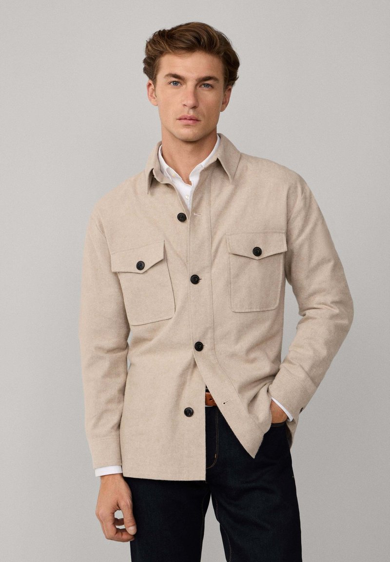 Camisa chaqueta de botones beige con dos bolsillos en el pecho, botones negros y una textura suave, combinada con jeans oscuros. Diseño y ajuste sencillos.