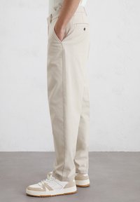 Beige Baumwollhosen im geraden Schnitt, mit Seiten- und einer Gesäßtasche mit Knopfdetail, getragen mit weißen und braunen Sneakers.